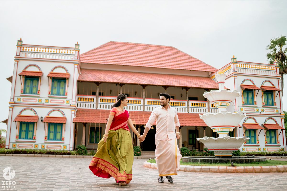 Couple in traditional South Indian attire holding hands outside a grand colonial-style building with red roof.