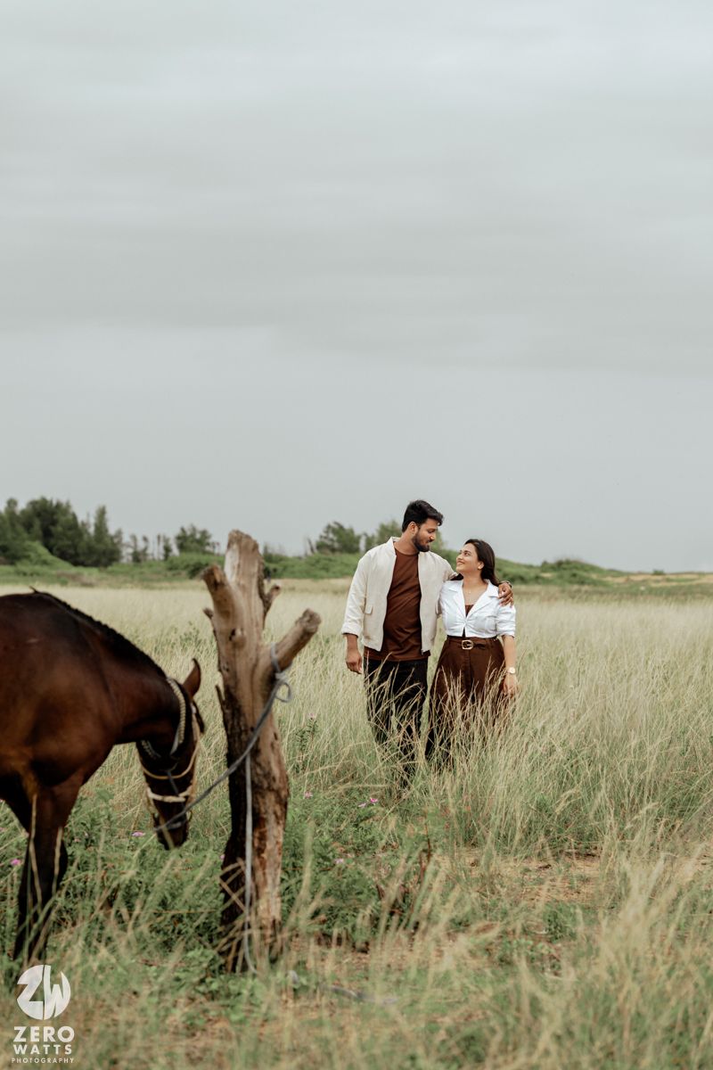 A romantic countryside photoshoot with a couple and their brown horse in a tall grass meadow under cloudy skies.