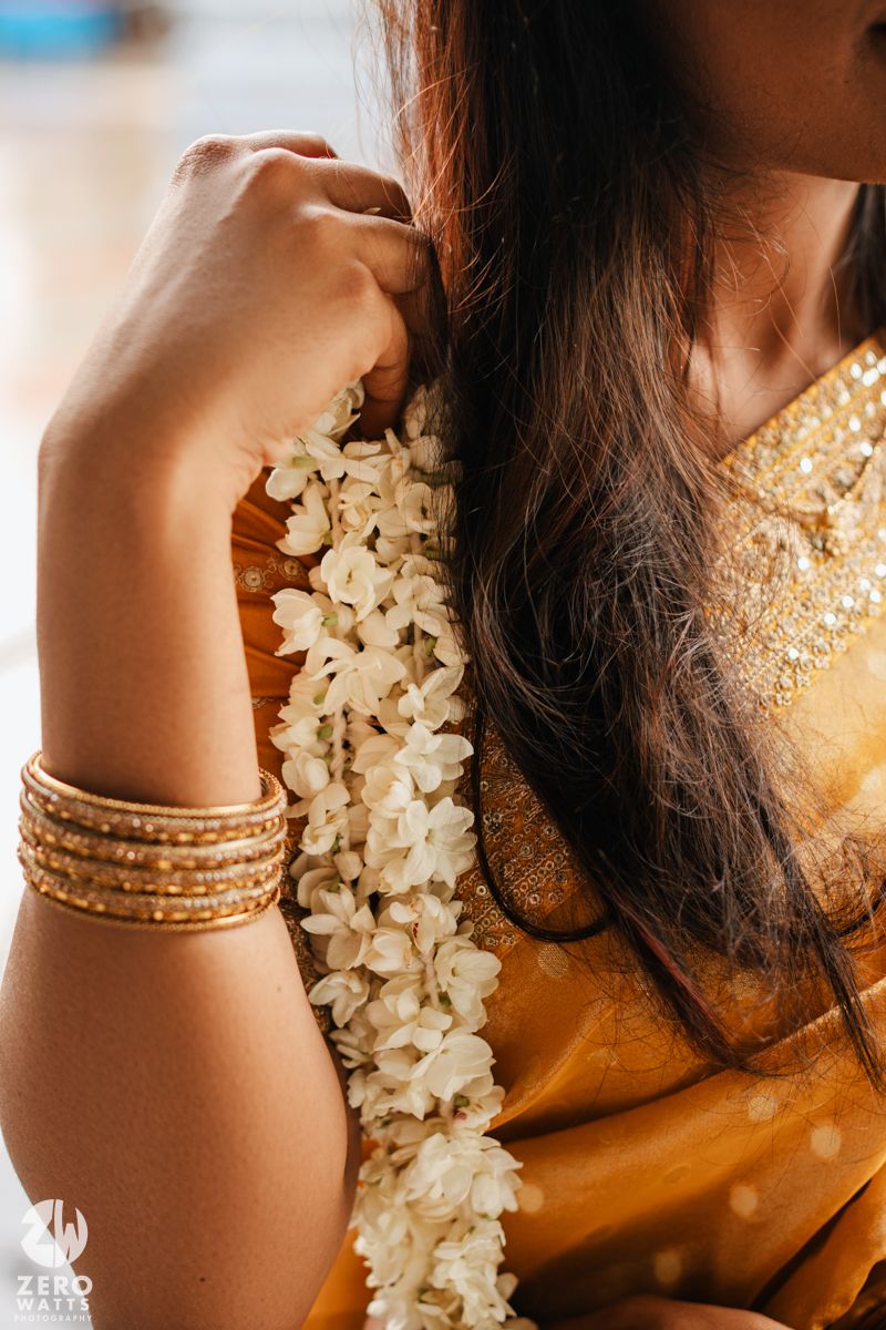 Close-up of gold bangles and white jasmine flowers adorning a wrist against a warm golden background.