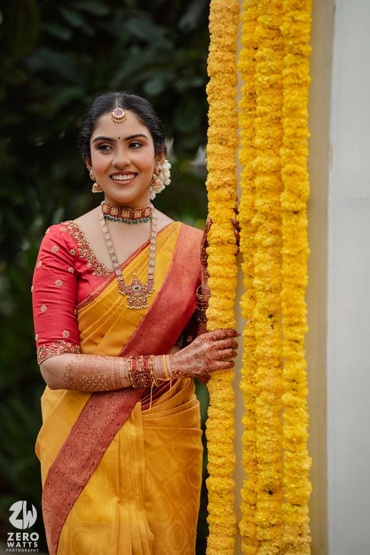 Traditional South Indian bride in yellow silk saree and red blouse poses beside marigold flower decorations.