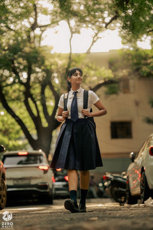 Person in sunglasses and dark vest poses casually against outdoor urban backdrop with trees and parked cars.