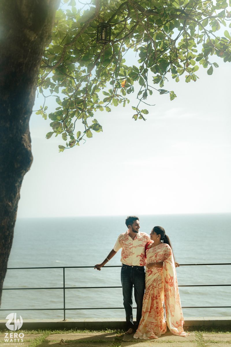 A couple in traditional attire share a romantic moment by the ocean railing during sunset.
