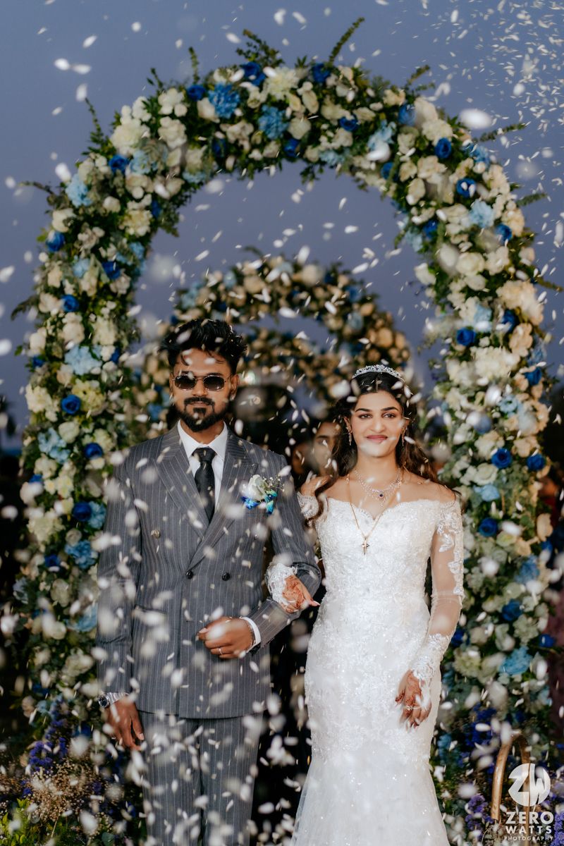 A floral wedding arch decorated with white and blue flowers frames a couple during their celebration while confetti falls.