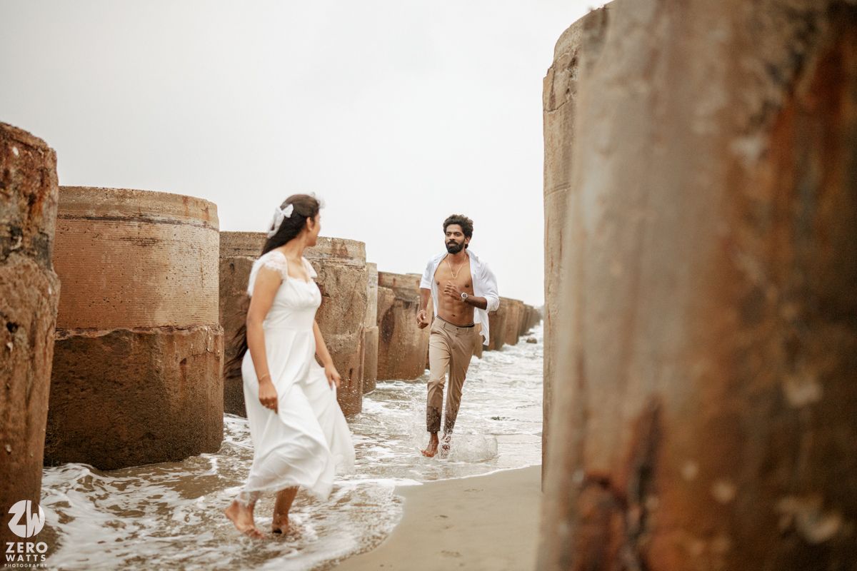 Couple walking between rustic concrete pillars on a beach, woman in white dress and man in casual attire.