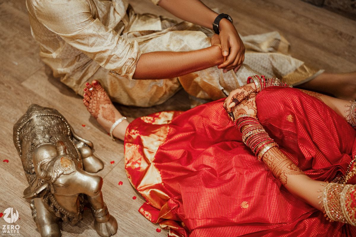 Indian bride and groom seated on floor with red rose petals and brass Ganesha idol during wedding ritual.