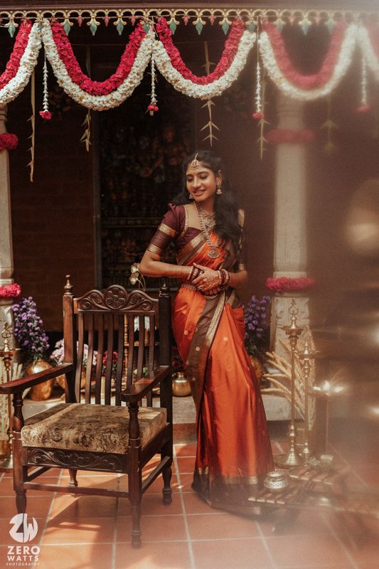 Bride in orange Kanjivaram silk saree with gold jewelry poses at decorated South Indian wedding venue.