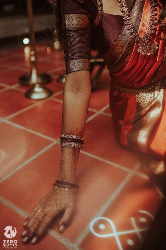 Close-up of a South Indian bride's arm adorned with silver bangles, mehndi, and a silk saree beside a kolam.