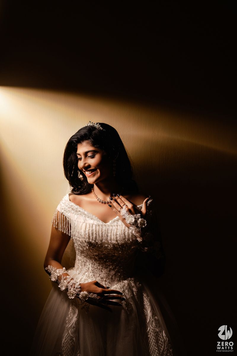 Bride in white beaded gown with tiara smiling under dramatic golden spotlight in dark studio setting.