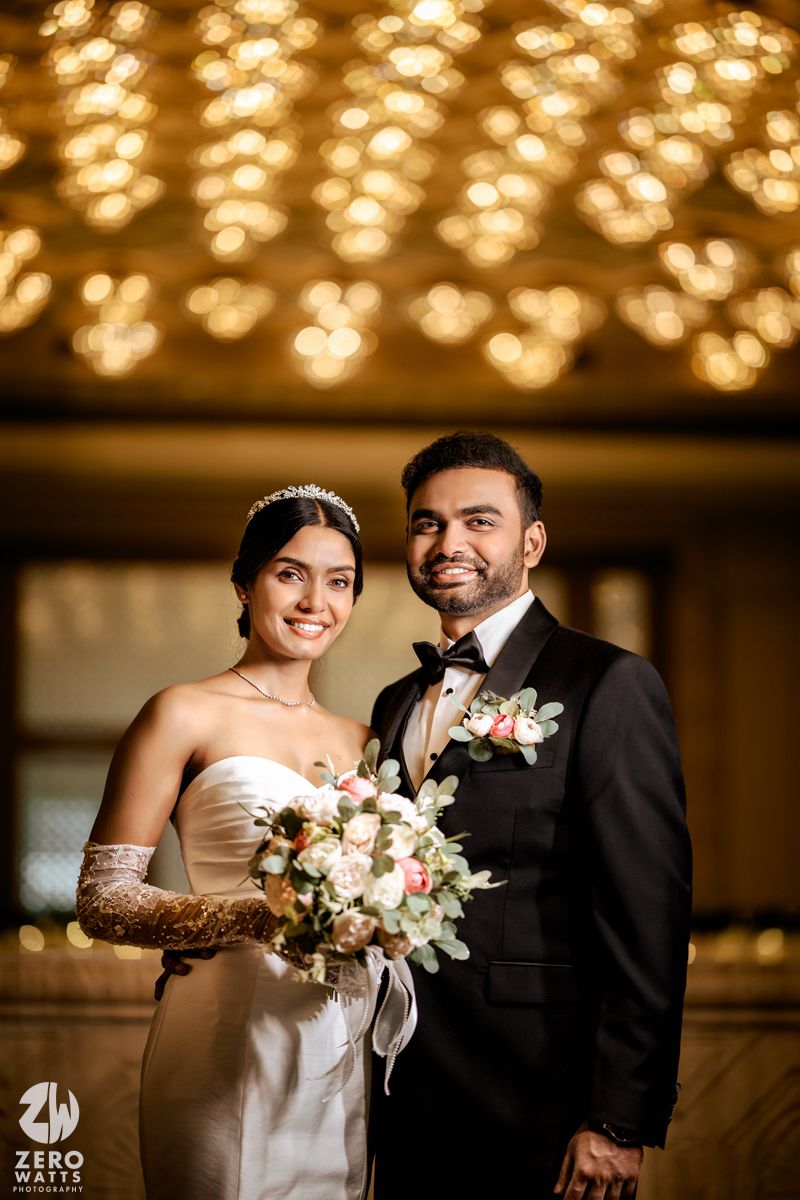 Newlyweds posing under golden bokeh lights, bride holding floral bouquet.