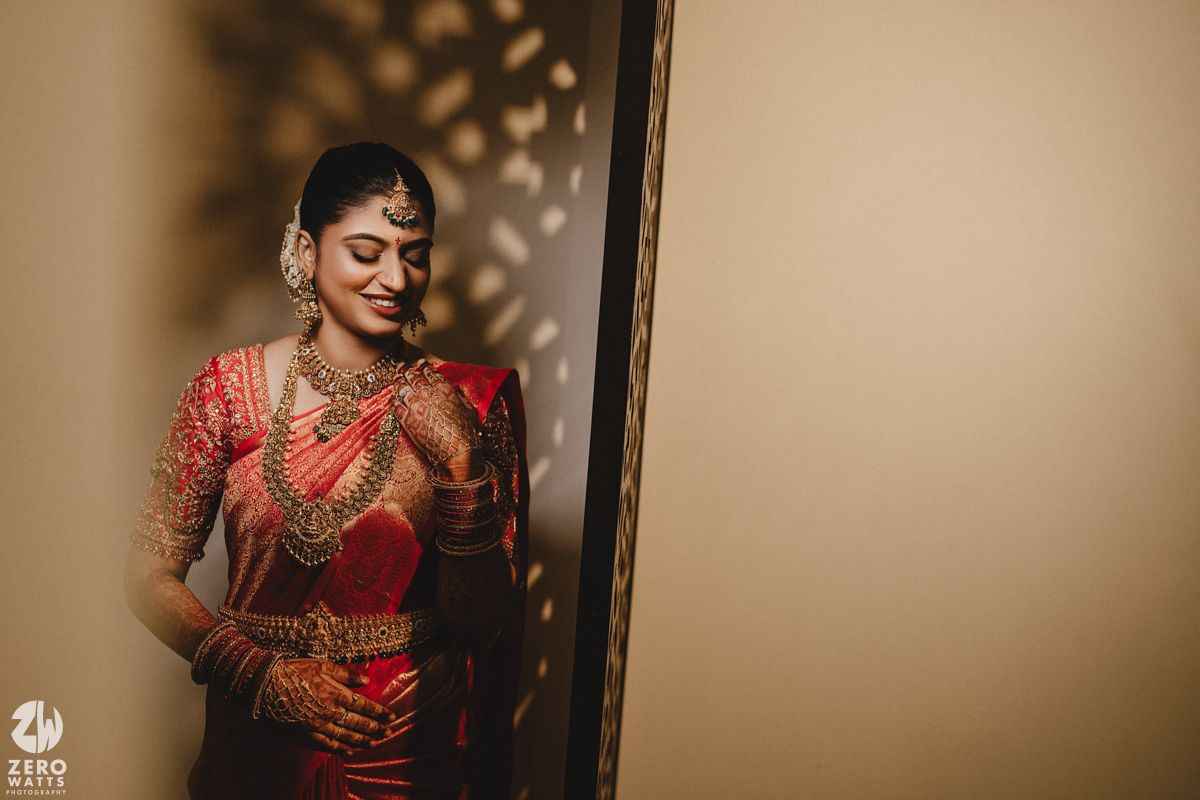 A bride in a traditional red and gold Indian wedding saree stands elegantly near a doorway in soft lighting.