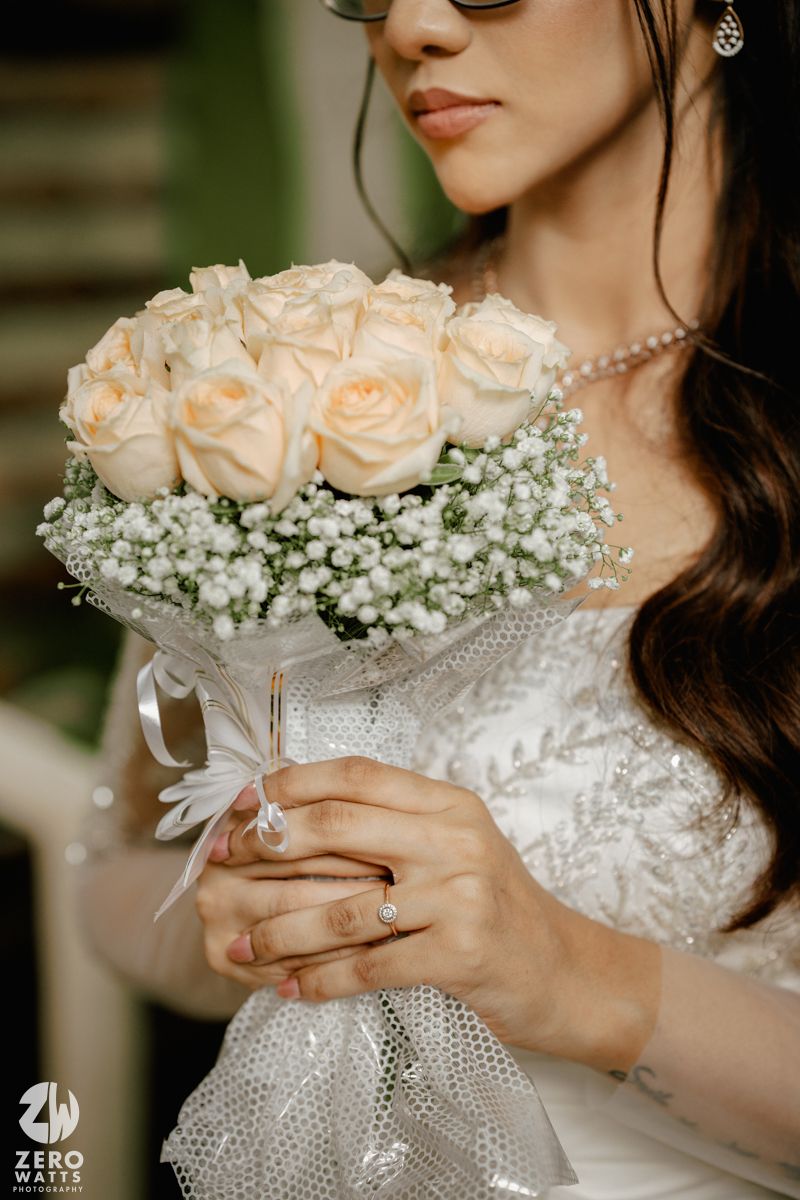 A delicate bridal bouquet of peach roses and baby's breath wrapped in tulle, held by hands wearing an engagement ring.