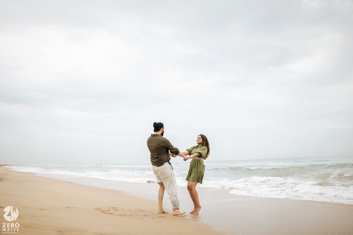 A couple walks hand in hand along a misty beach shoreline during an overcast day.