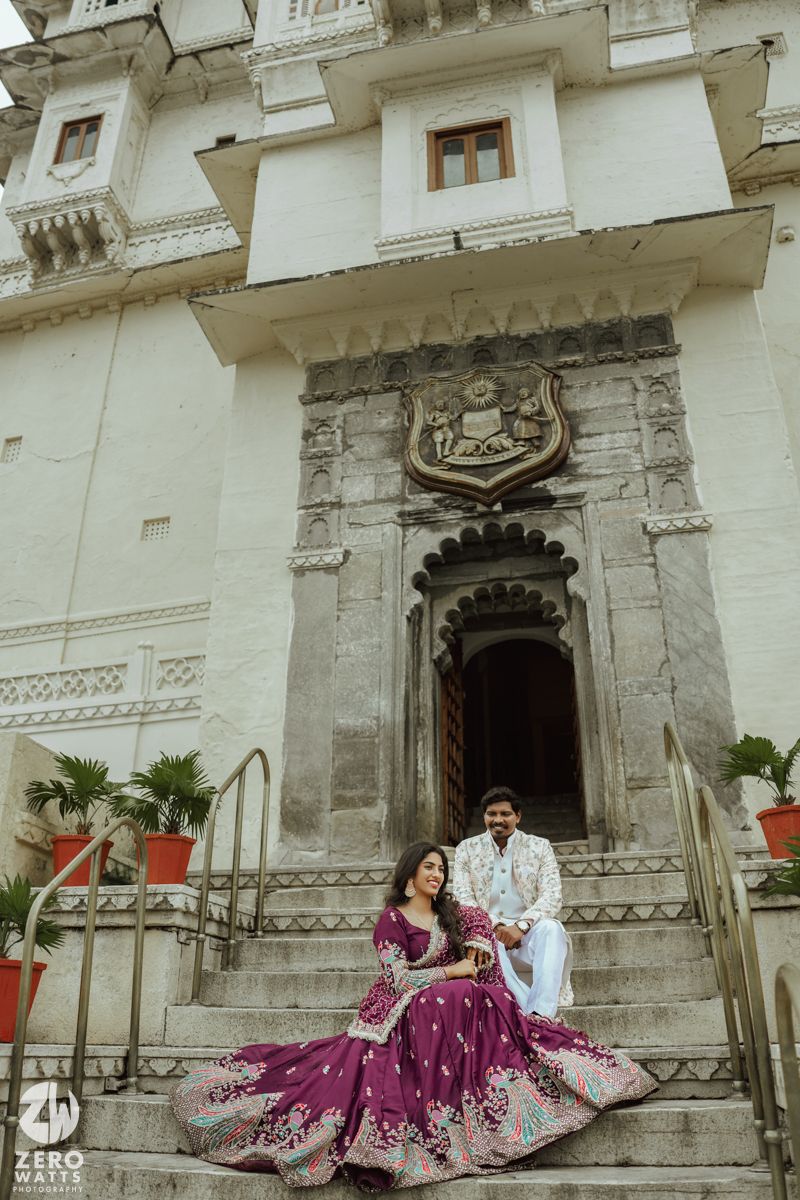 A couple in traditional Indian attire poses on ornate palace steps with white walls and decorative architecture.