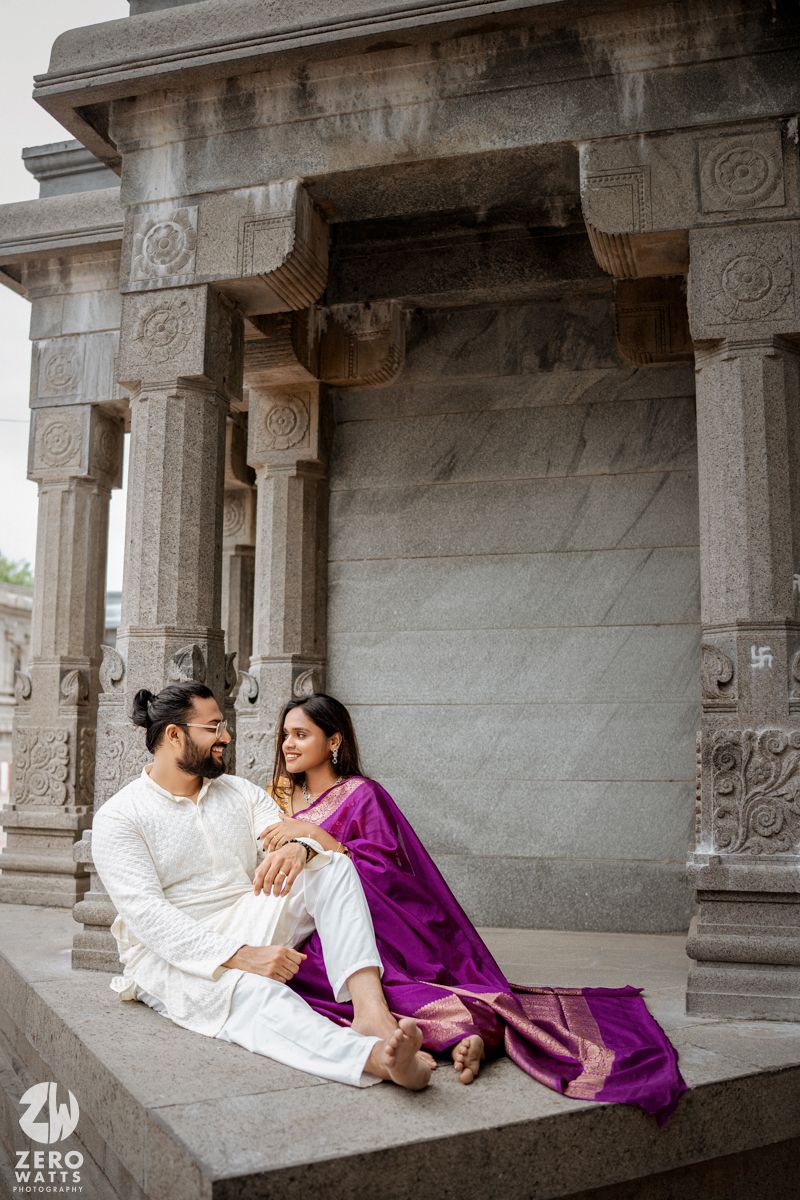 A couple in traditional Indian attire lounges in a temple corridor with ornate stone pillars and architecture.