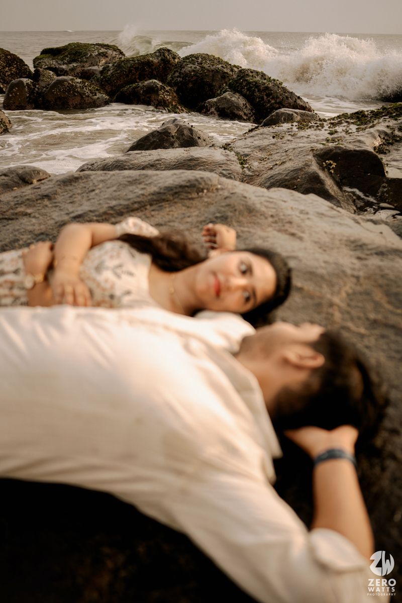 A romantic couple lying on dark coastal rocks in white clothing at sunset.