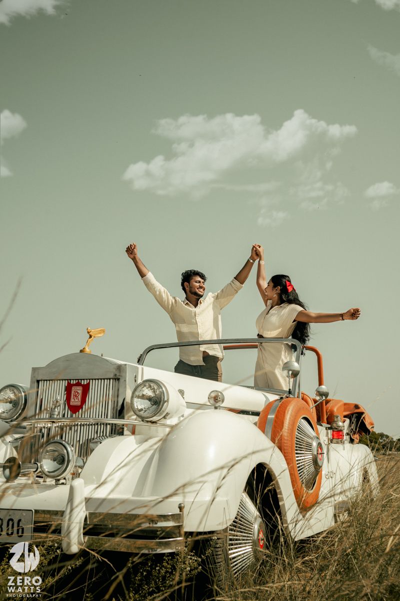 Joyful couple celebrating with raised arms while standing in a vintage white car in a field.