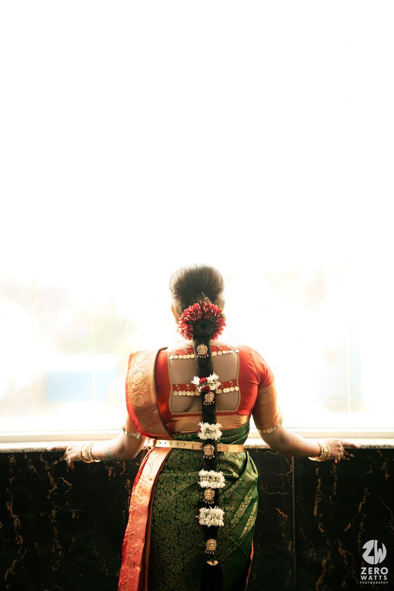 South Indian bride in green and red silk saree with floral hair accessories and gold jewelry, viewed from behind.