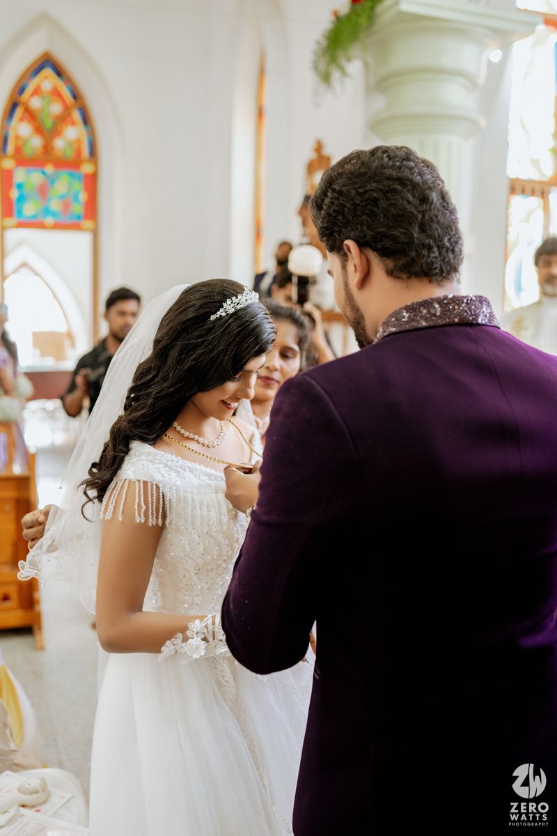 Bride in white lace gown and veil receiving necklace from groom in purple suit during church wedding ceremony.