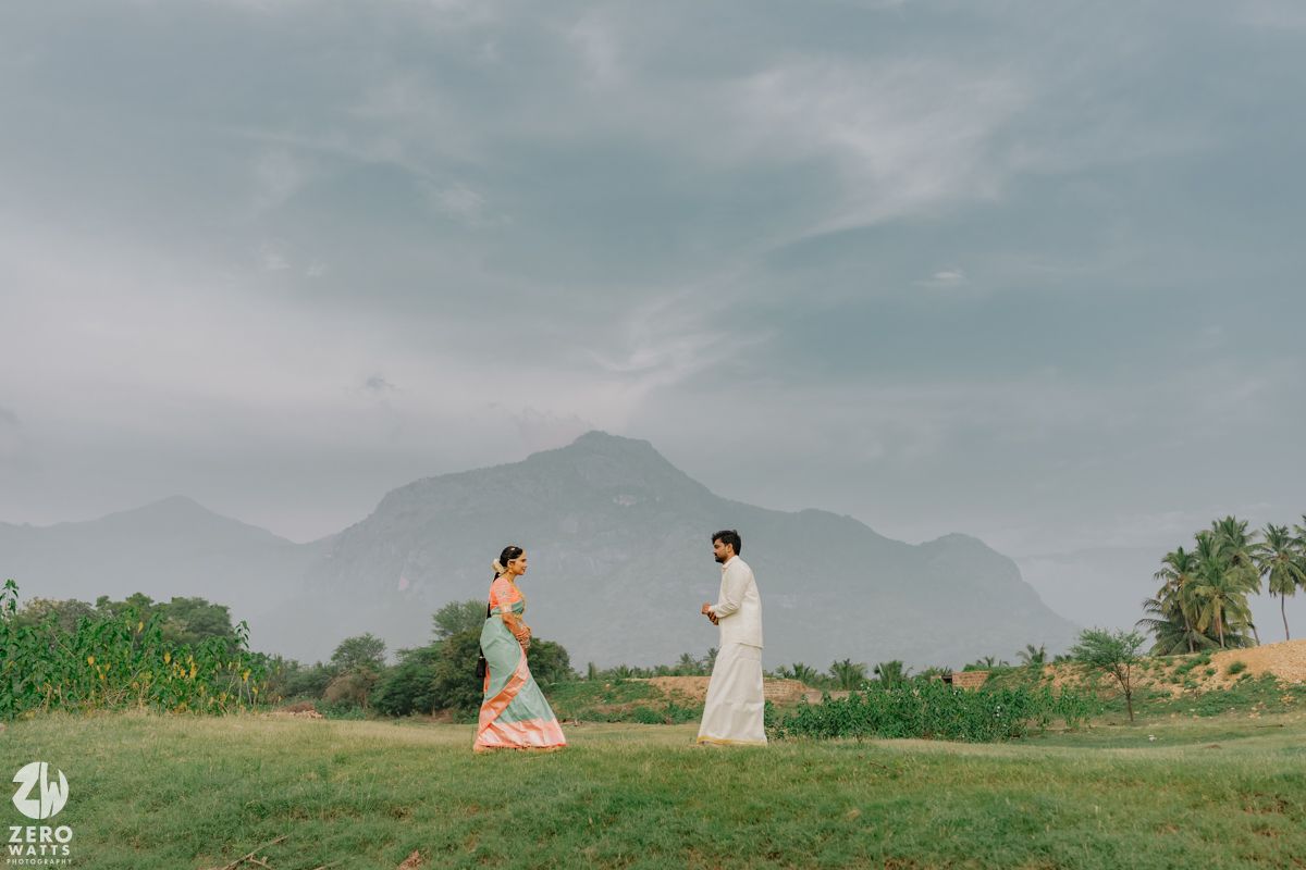 Indian bride and groom walking separately on green field with mountain backdrop.
