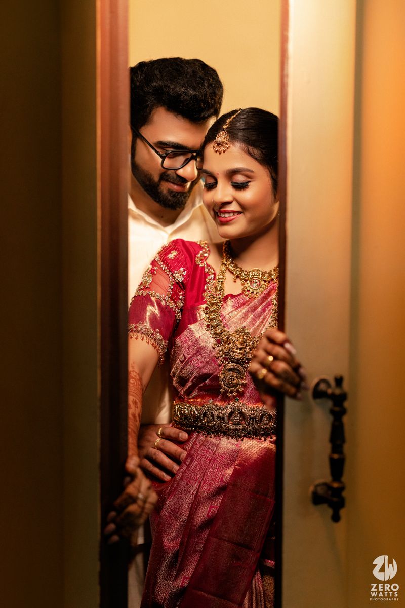 South Indian bride in pink silk saree with gold jewelry embraced by groom in white during wedding photoshoot.