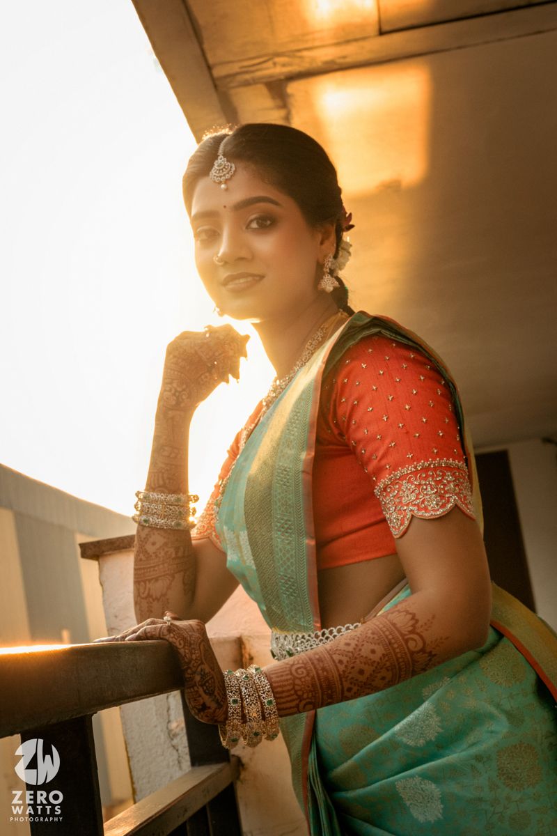 Indian bride in teal saree and orange blouse with mehndi, posing at golden hour with traditional bridal jewelry.