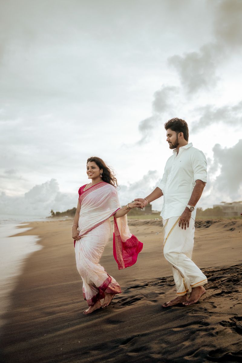 Two people in white and pink attire run playfully across sandy dunes under cloudy skies.
