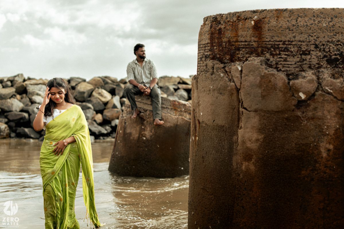 A couple explores a rocky coastal area with ancient stone structures in the background.