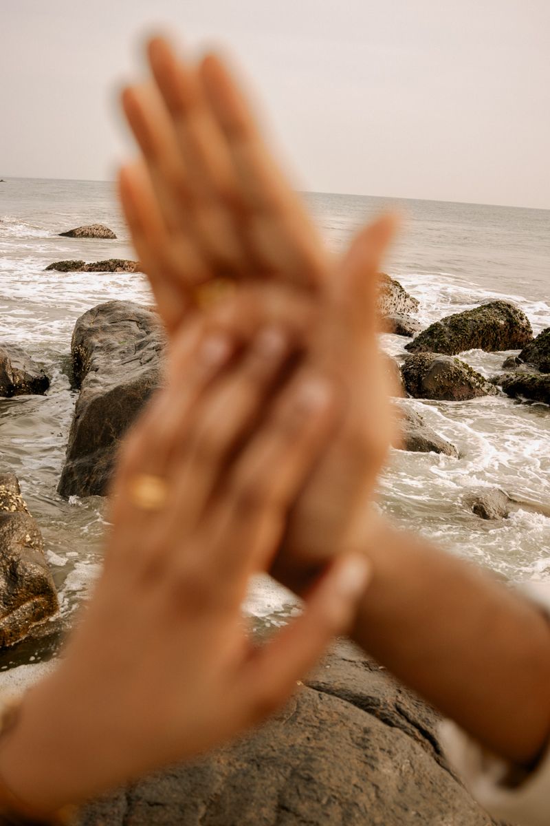 Close up of hands with wedding rings against a blurred ocean and rocky beach backdrop.