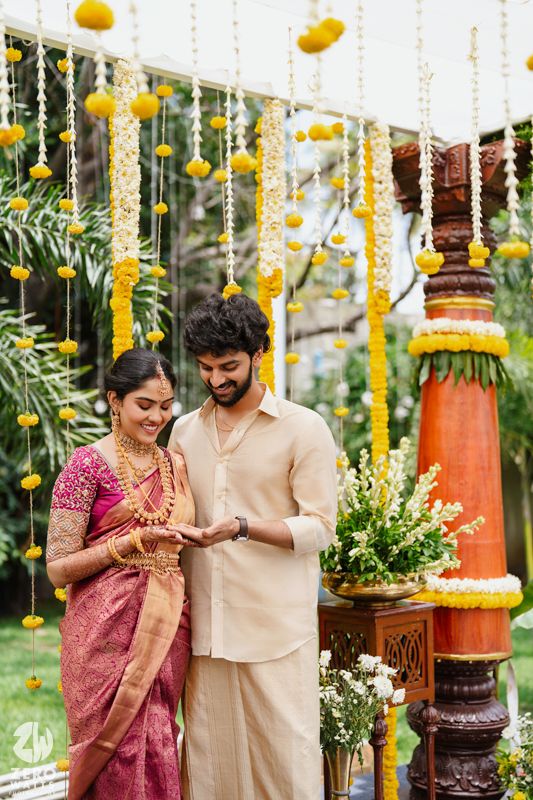 Couple in traditional Indian wedding attire under hanging yellow marigold decorations and wooden decor.