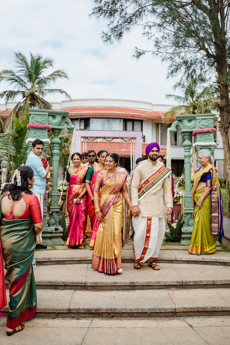 Traditional Indian wedding ceremony guests gather on ornate steps with palm trees and architecture in the background.