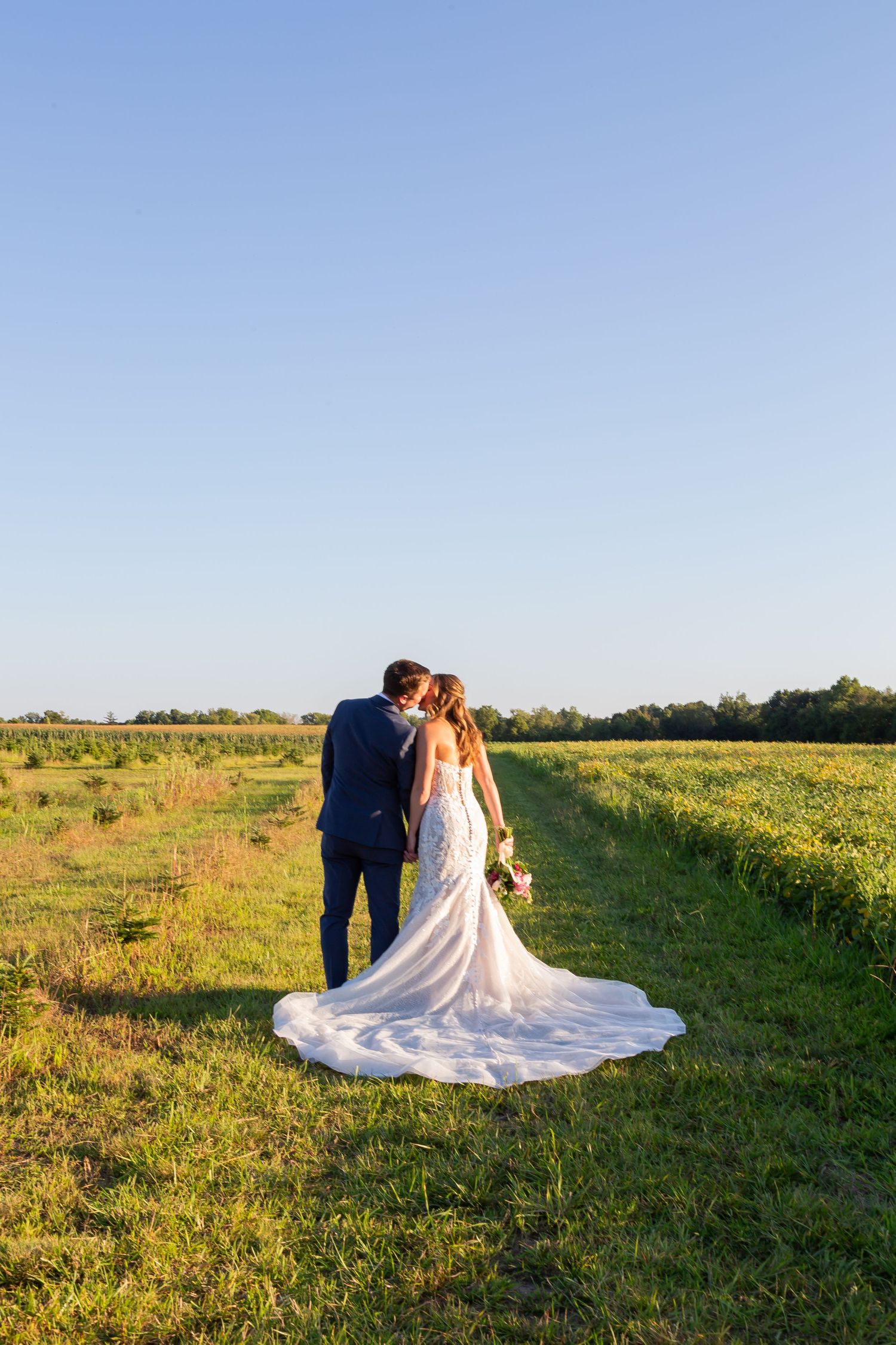 A couple embraces in a sunlit meadow during golden hour, the bride's white lace dress trailing beautifully across green grass.