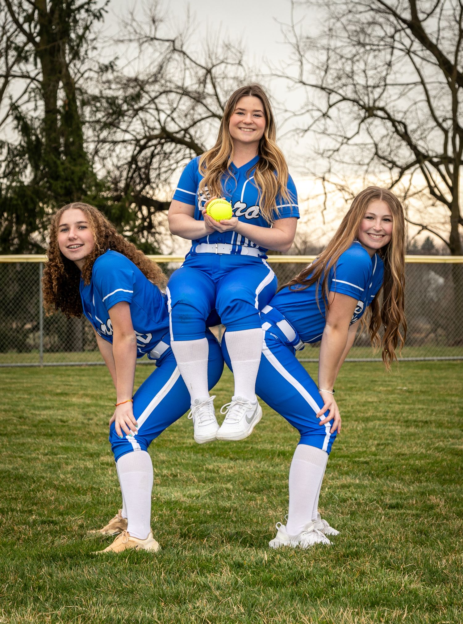 Three softball players in blue uniforms create a playful pyramid pose on the softball field.