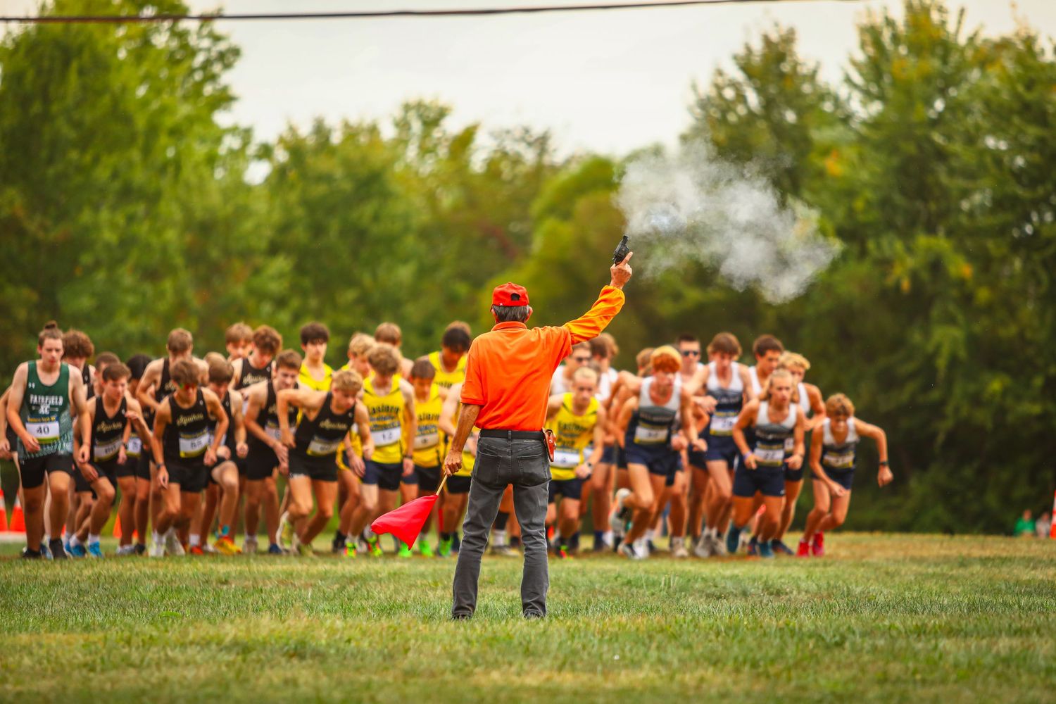 A race official in orange signals the start of a cross country meet as runners begin on a grassy course.