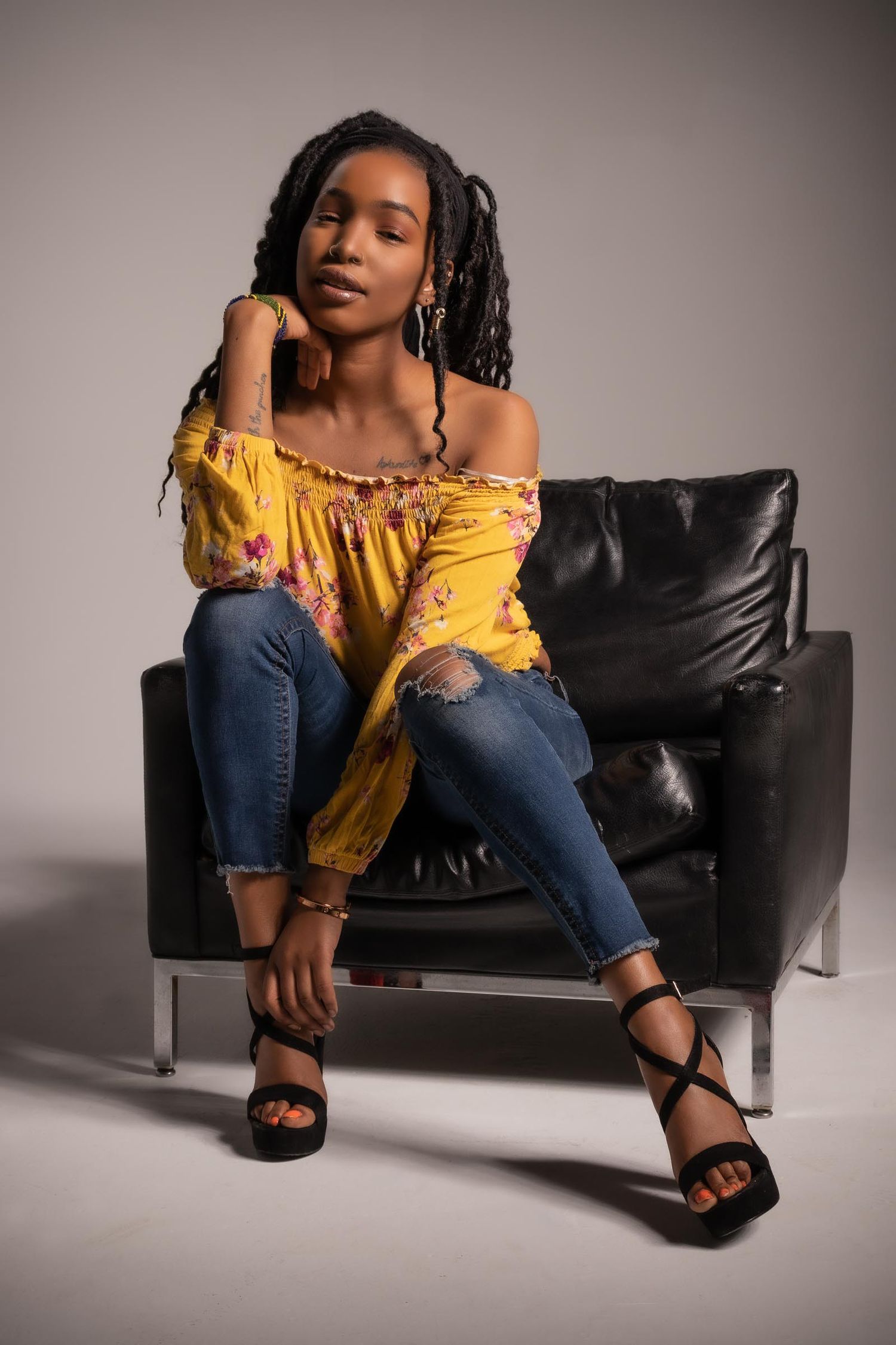Artistic portrait of black female model in yellow top and jeans on leather chair.