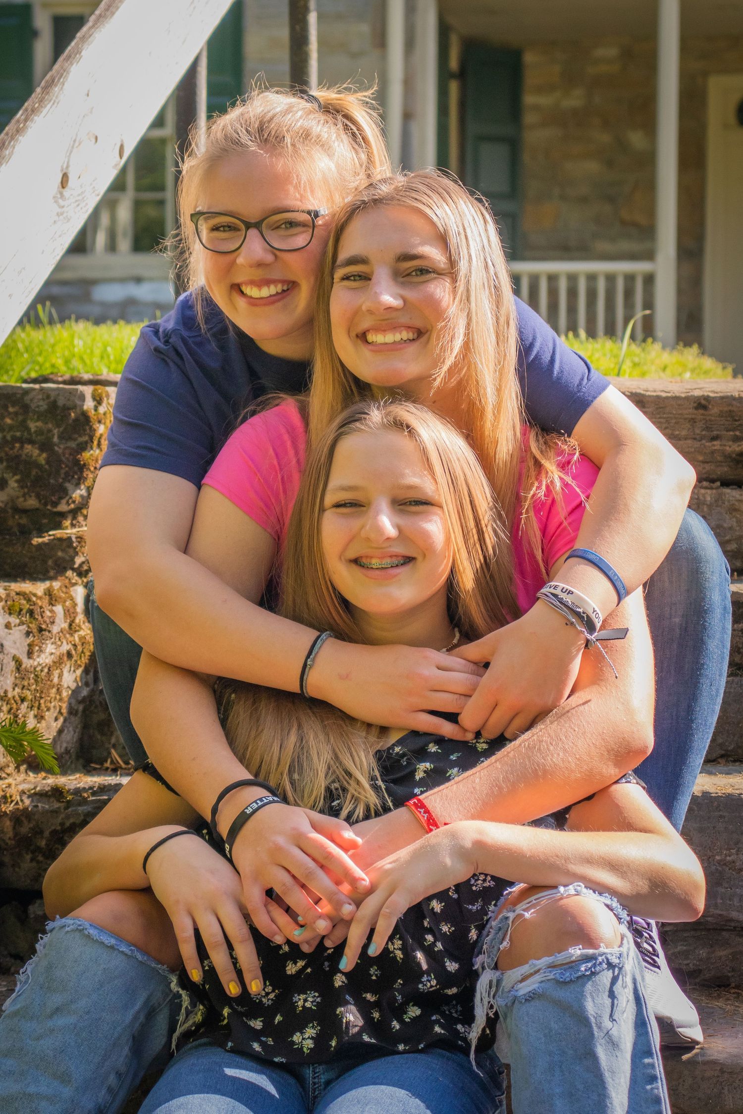 Three sisters share warm embrace while sitting on outdoor stone steps in bright sunlight.