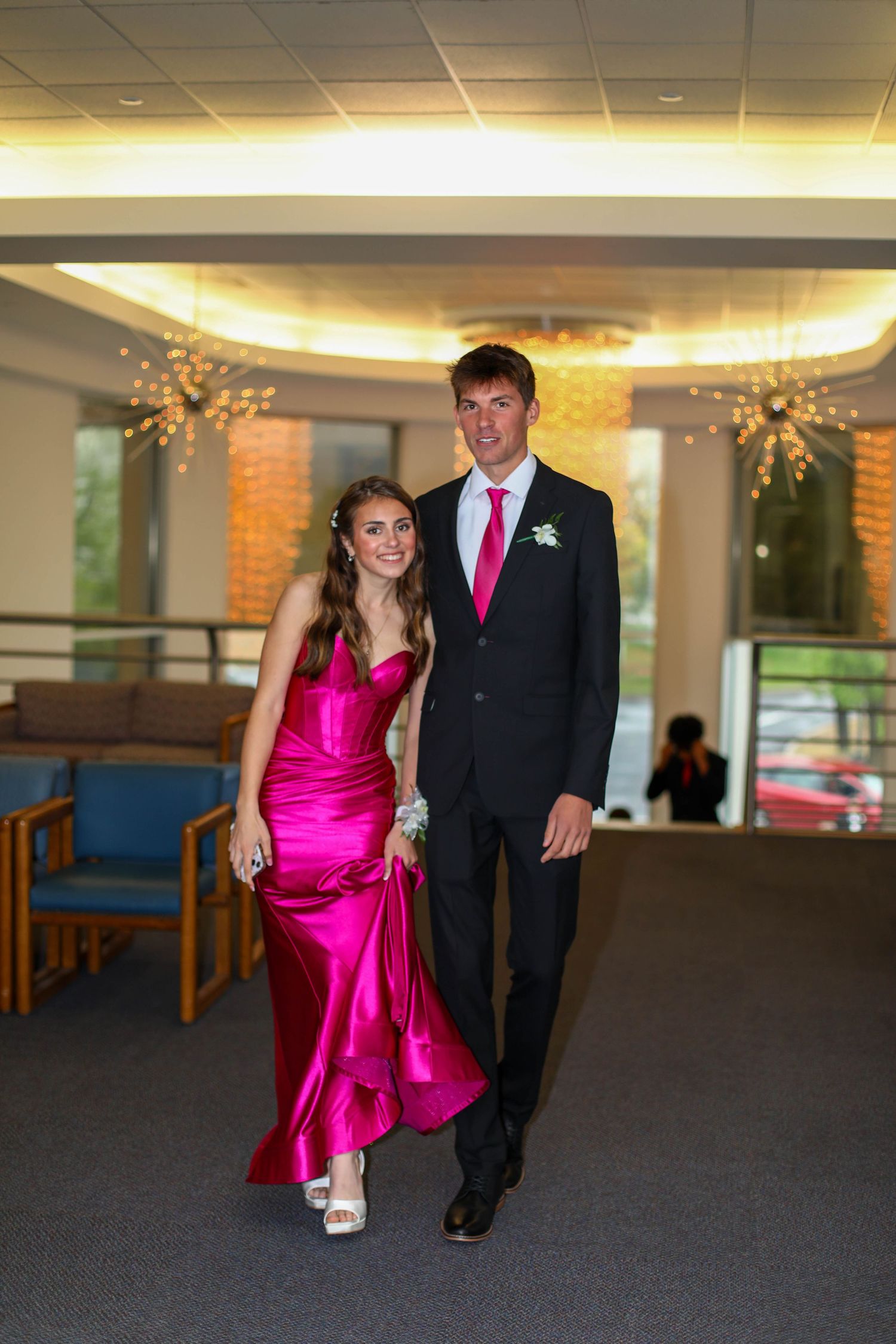 A couple in formal attire walks together, with the woman wearing a bright pink satin gown.