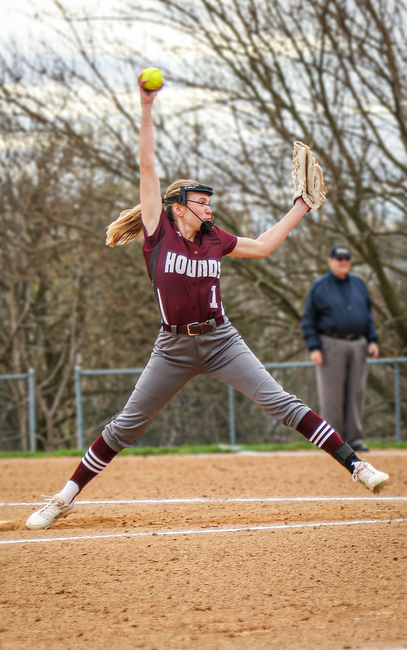 Softball pitcher in maroon uniform at peak of windmill pitching motion.