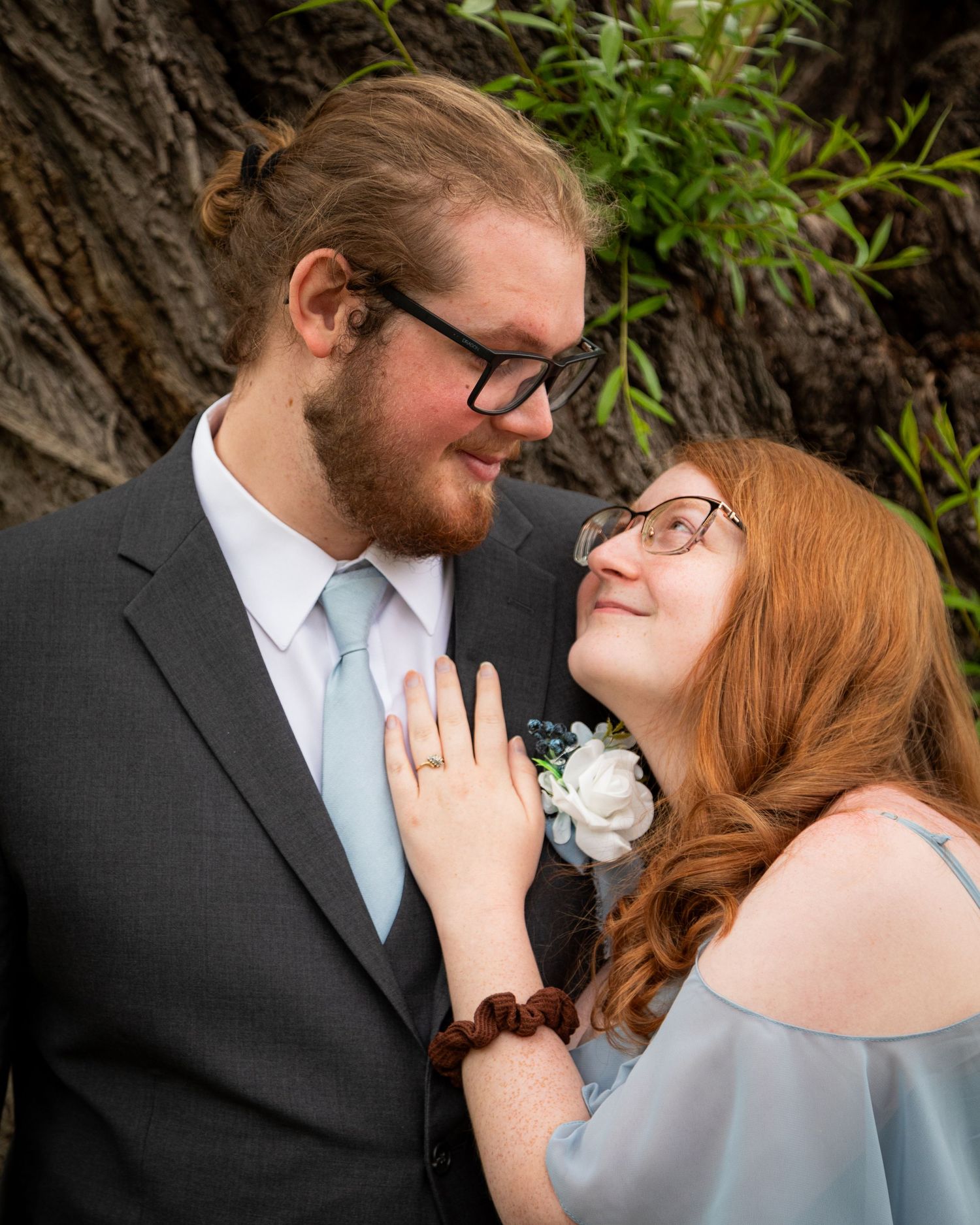 Romantic engagement photo of couple in formal attire by tree trunk in natural outdoor setting.