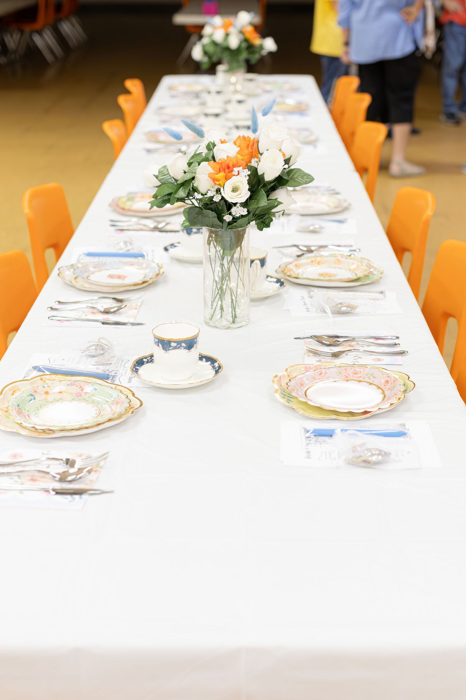 Elegant table setting with white tablecloth, orange chairs and floral centerpiece in glass vase with vintage china place settings.