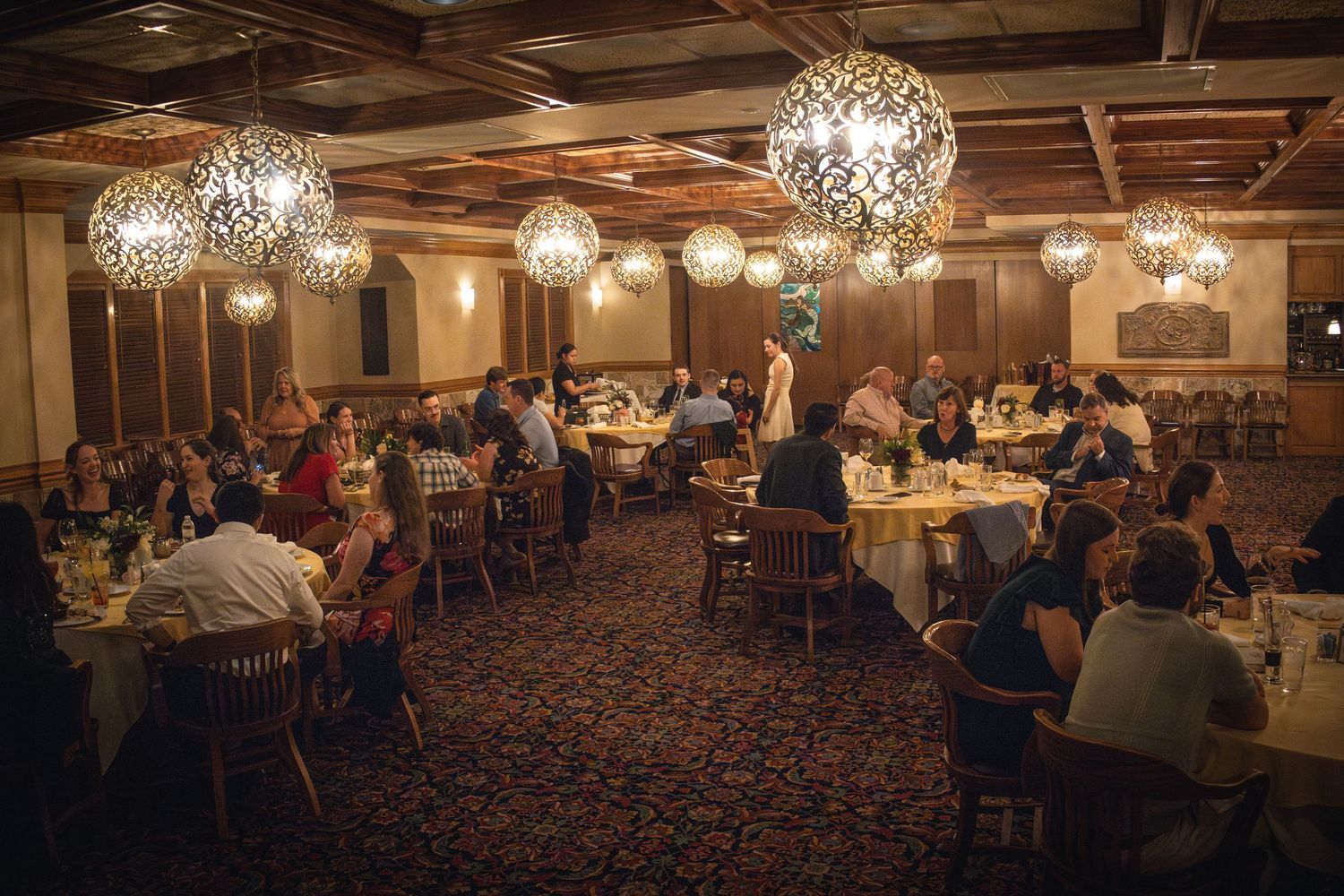 Elegant dining room with ornate crystal chandeliers illuminating guests seated at round tables on patterned carpet.