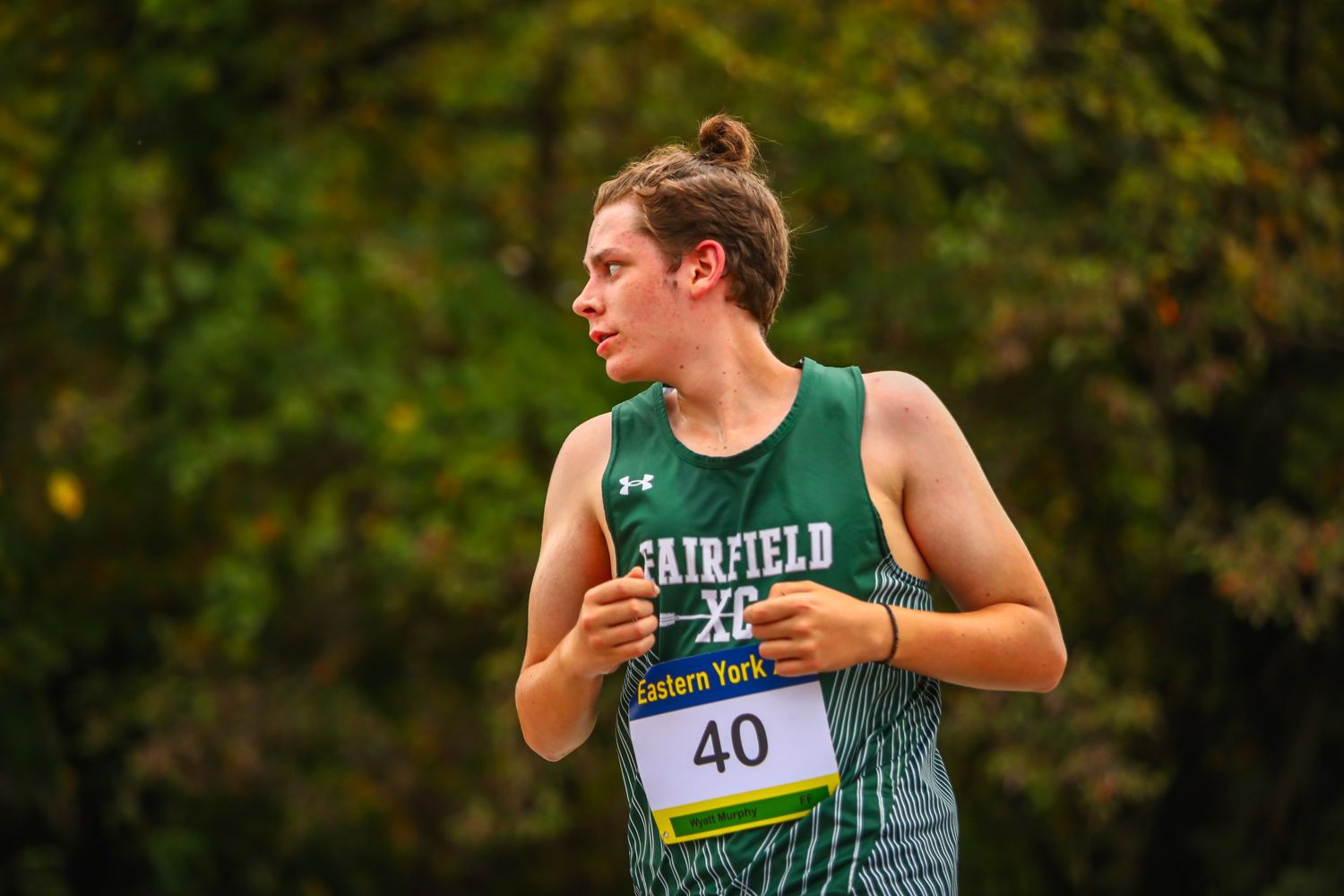 Cross country runner in green singlet competes on wooded course.