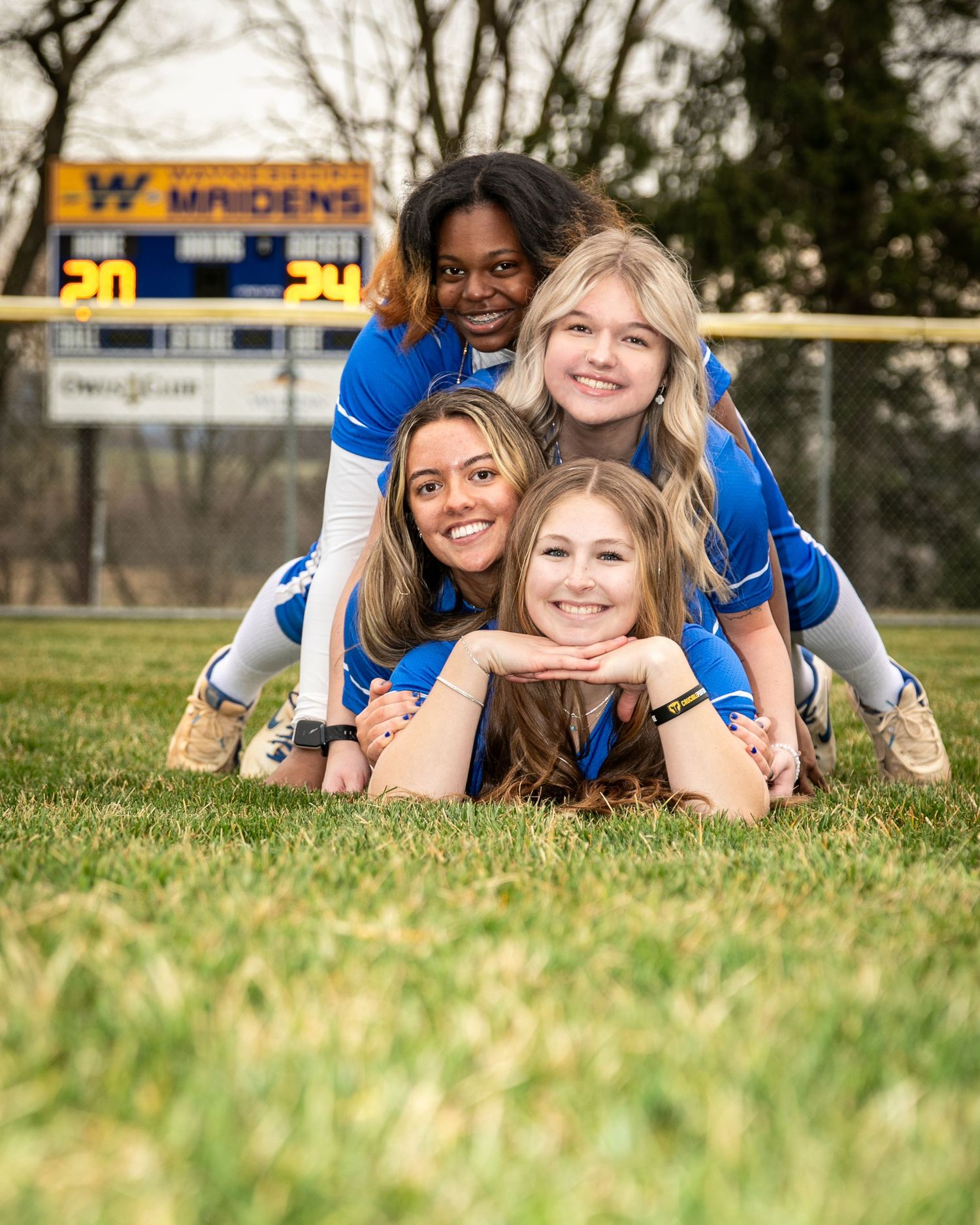 A group of softball teammates pose playfully stacked on each other on a field with scoreboard visible.