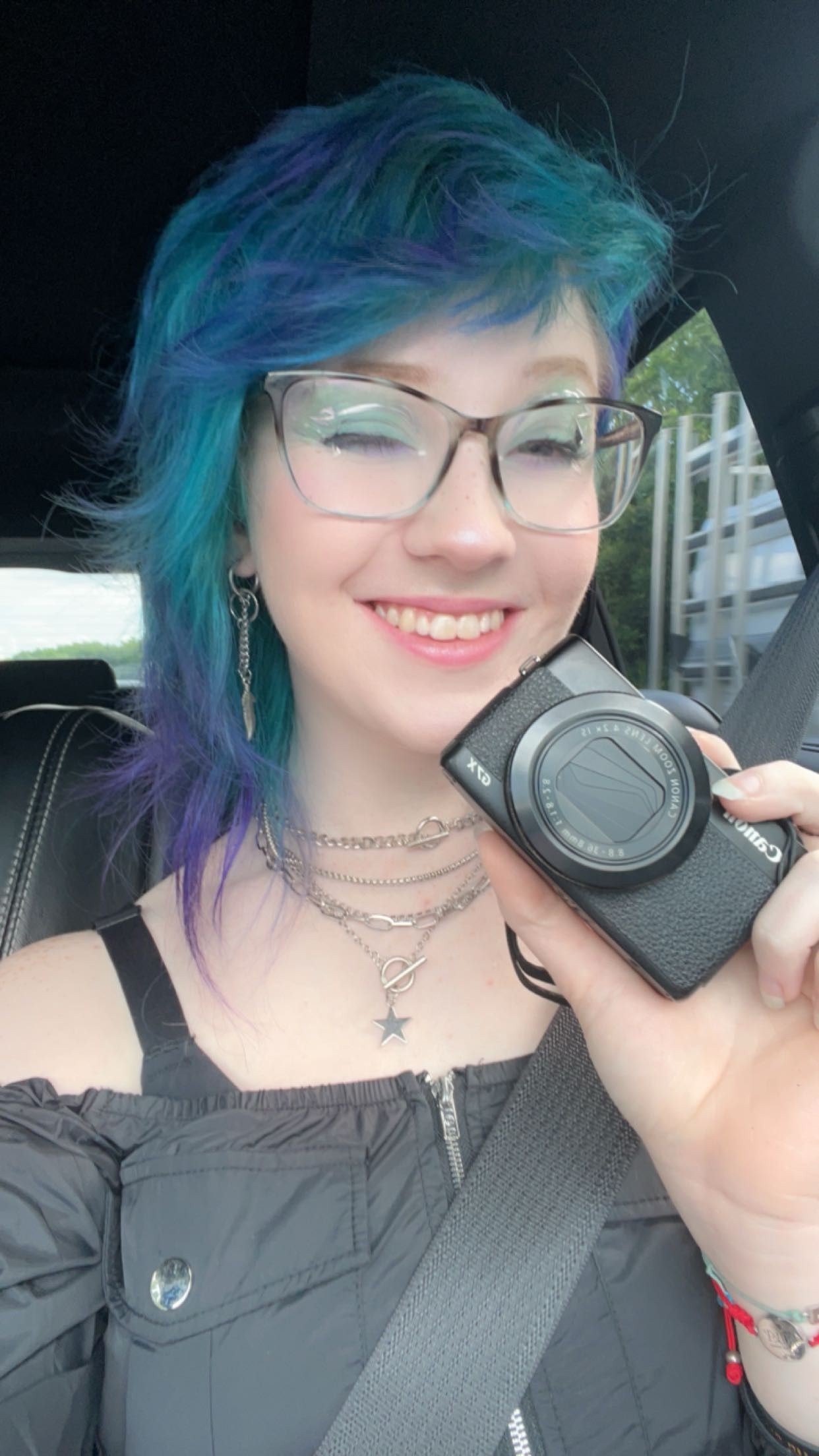 Person with vibrant blue-purple hair holding a camera and wearing multiple necklaces.