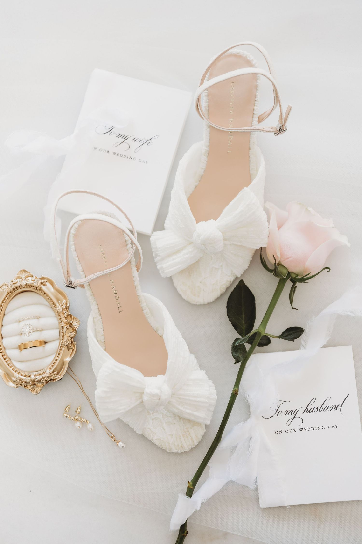 White bridal heels with bow detail beside a pink rose, wedding rings, and \u201cTo My Husband\u201d card on white fabric.