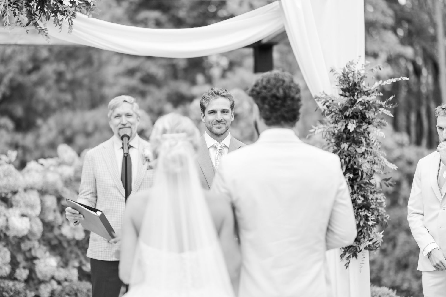 Black and white outdoor wedding ceremony with groom smiling at bride under floral arch, officiant holding book.