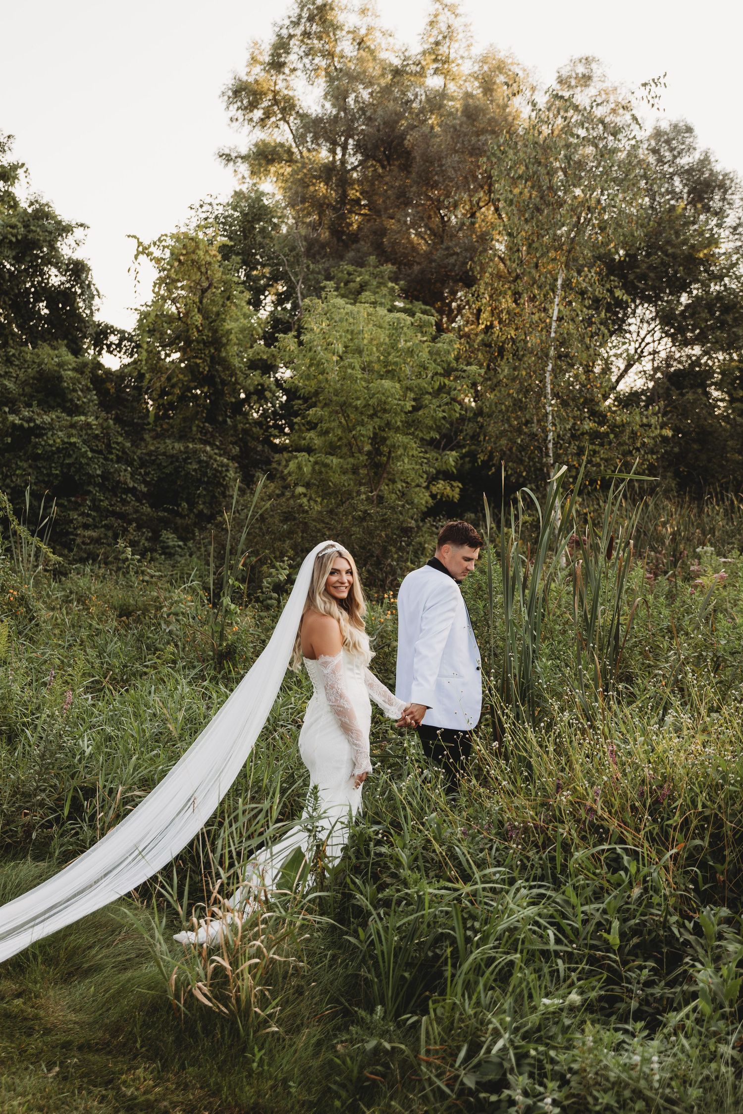 A bride in a flowing white gown and veil holds hands with a groom in a white suit amid lush greenery.