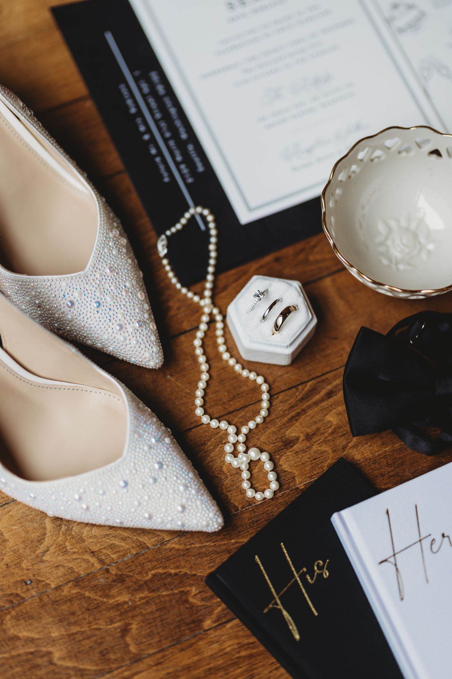Elegant wedding flat lay with pearl heels, pearl necklace, wedding rings, bow tie, and His and Hers vow books on wood.
