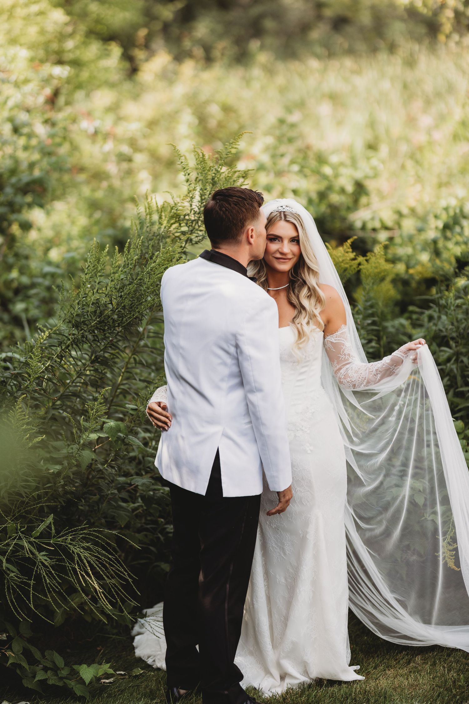 Groom in white tuxedo kissing bride in elegant white gown and veil outdoors amid lush green foliage.