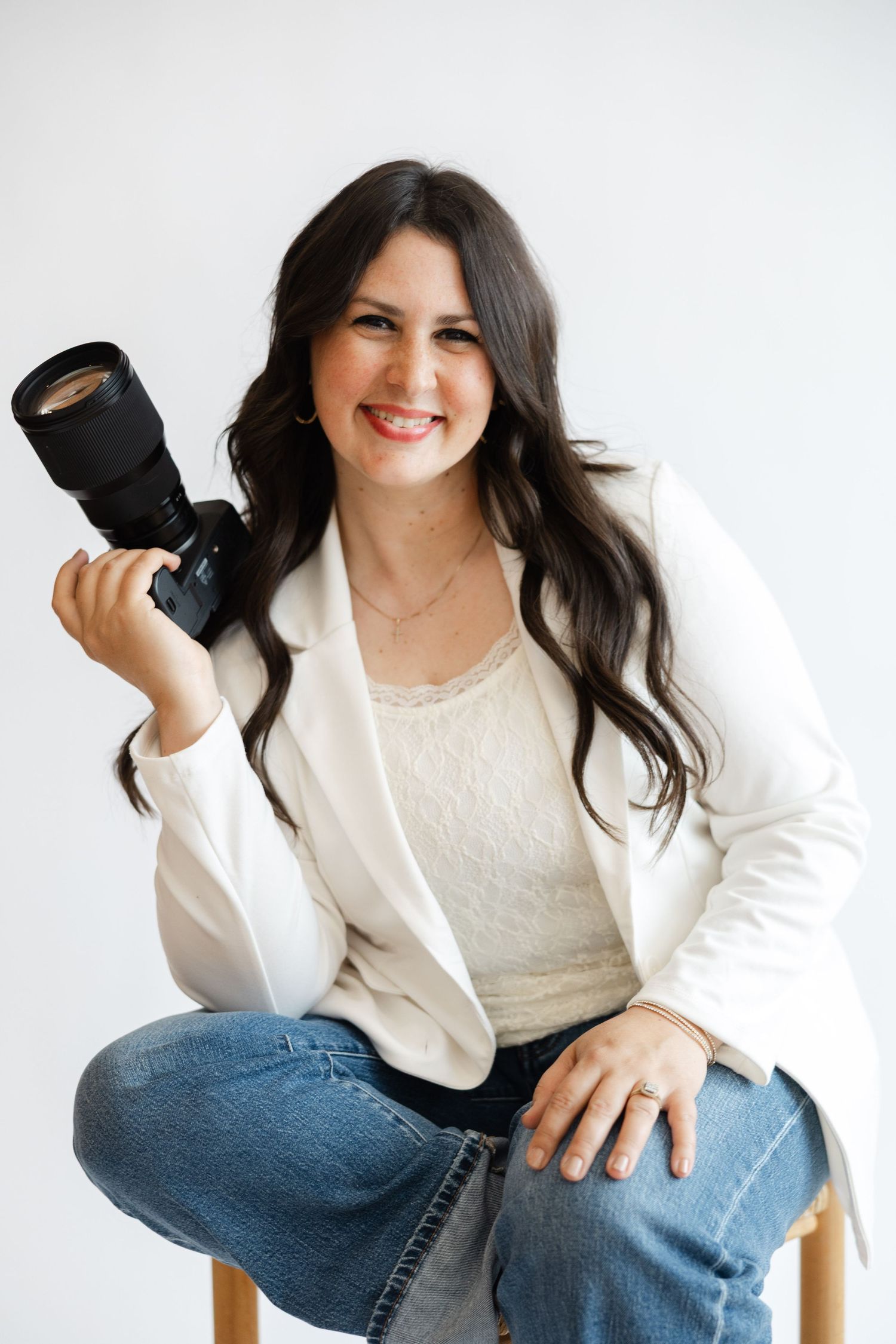 Smiling female photographer sitting on stool holding DSLR camera, wearing white blazer and jeans against white background.