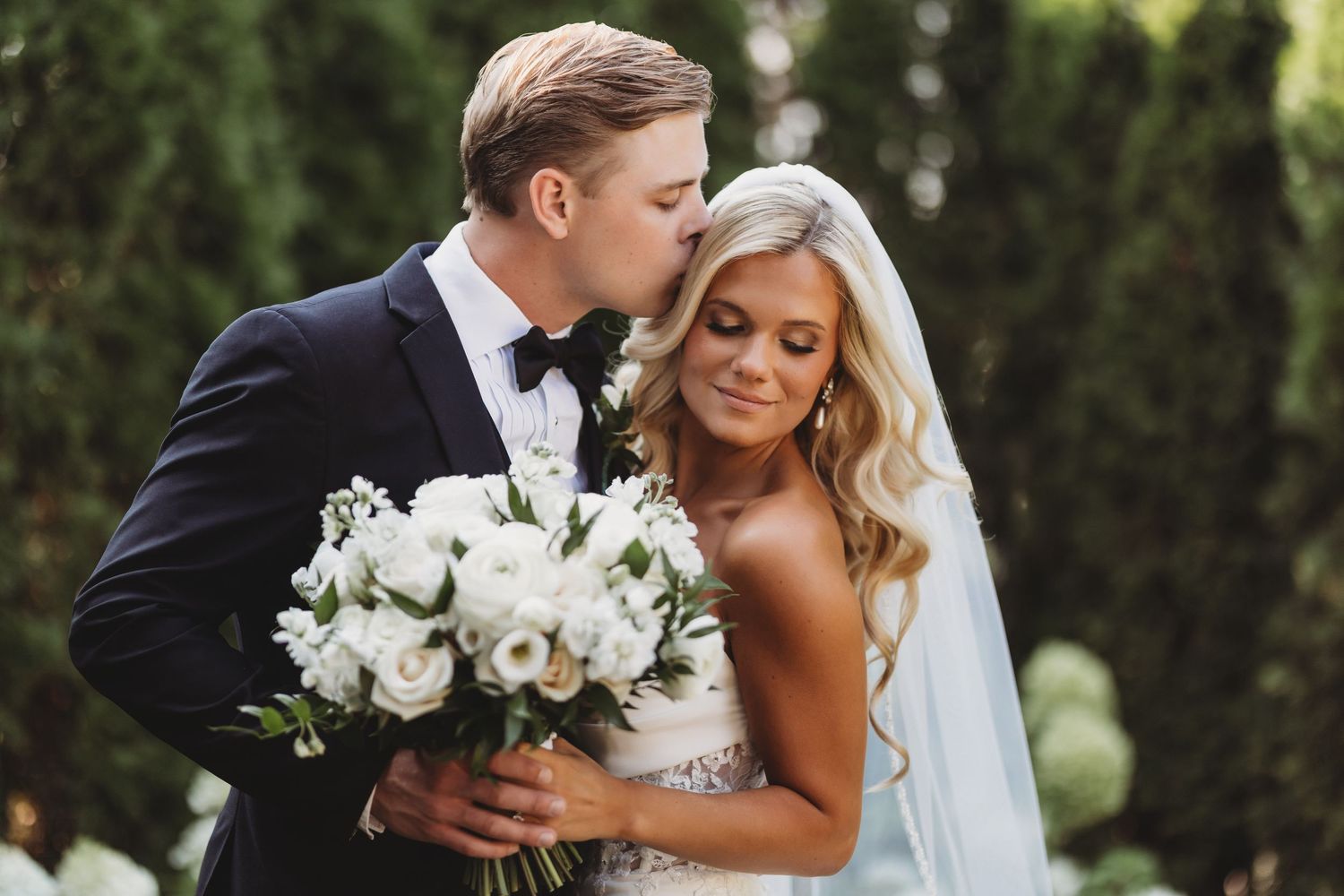 Bride and groom share a romantic moment, groom kissing bride's cheek as she holds a white floral bouquet outdoors.